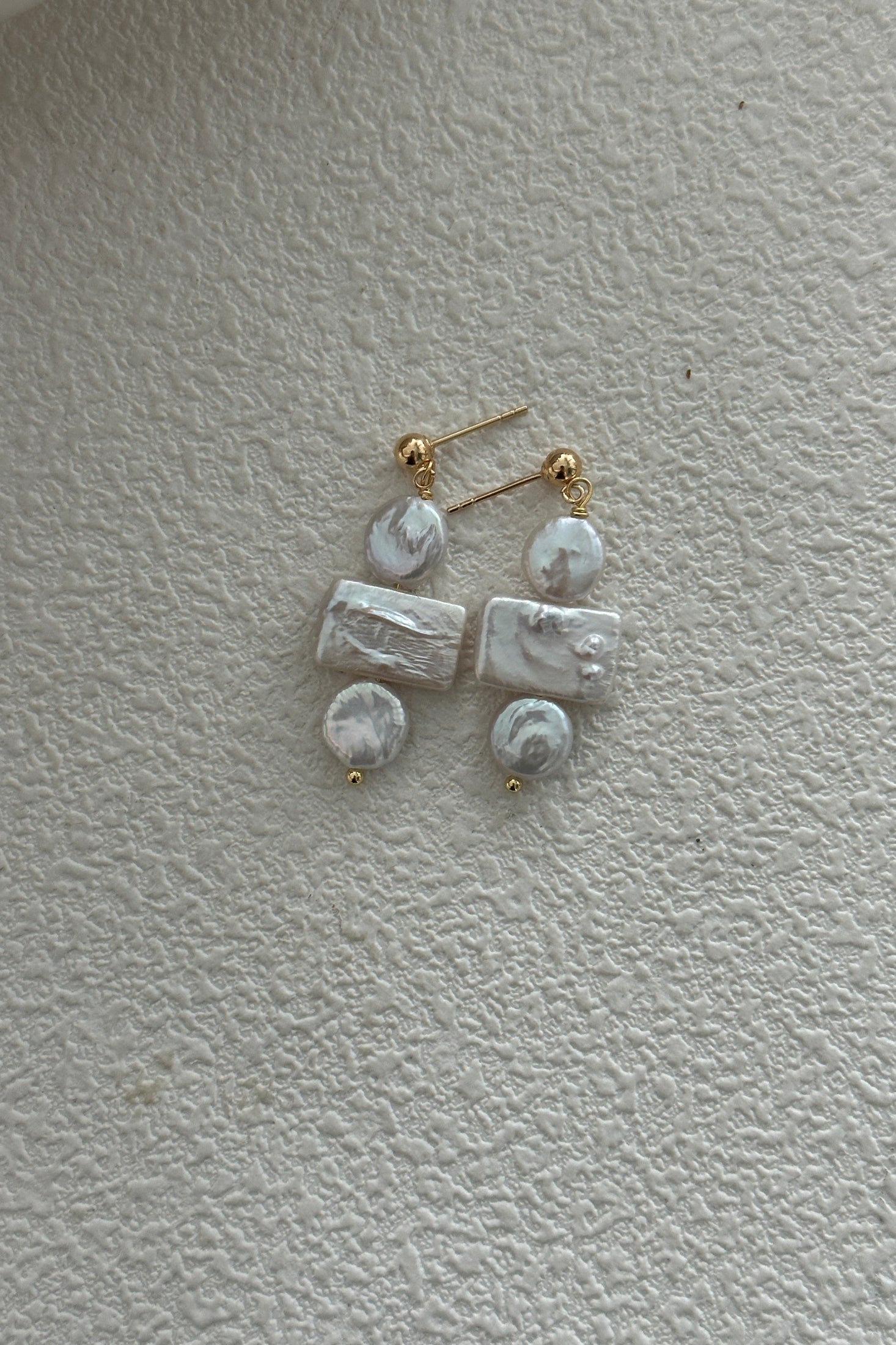 A pair of Siren Mode's 18K Gold Plated 925 Silver Baroque Pearl Drop Earrings, featuring exquisite studs at the top. Each earring showcases three white gemstones—one round, one rectangular, and one round—stacked vertically and connected by gold fixtures. The elegant Baroque Pearl design is accentuated on a textured white surface.