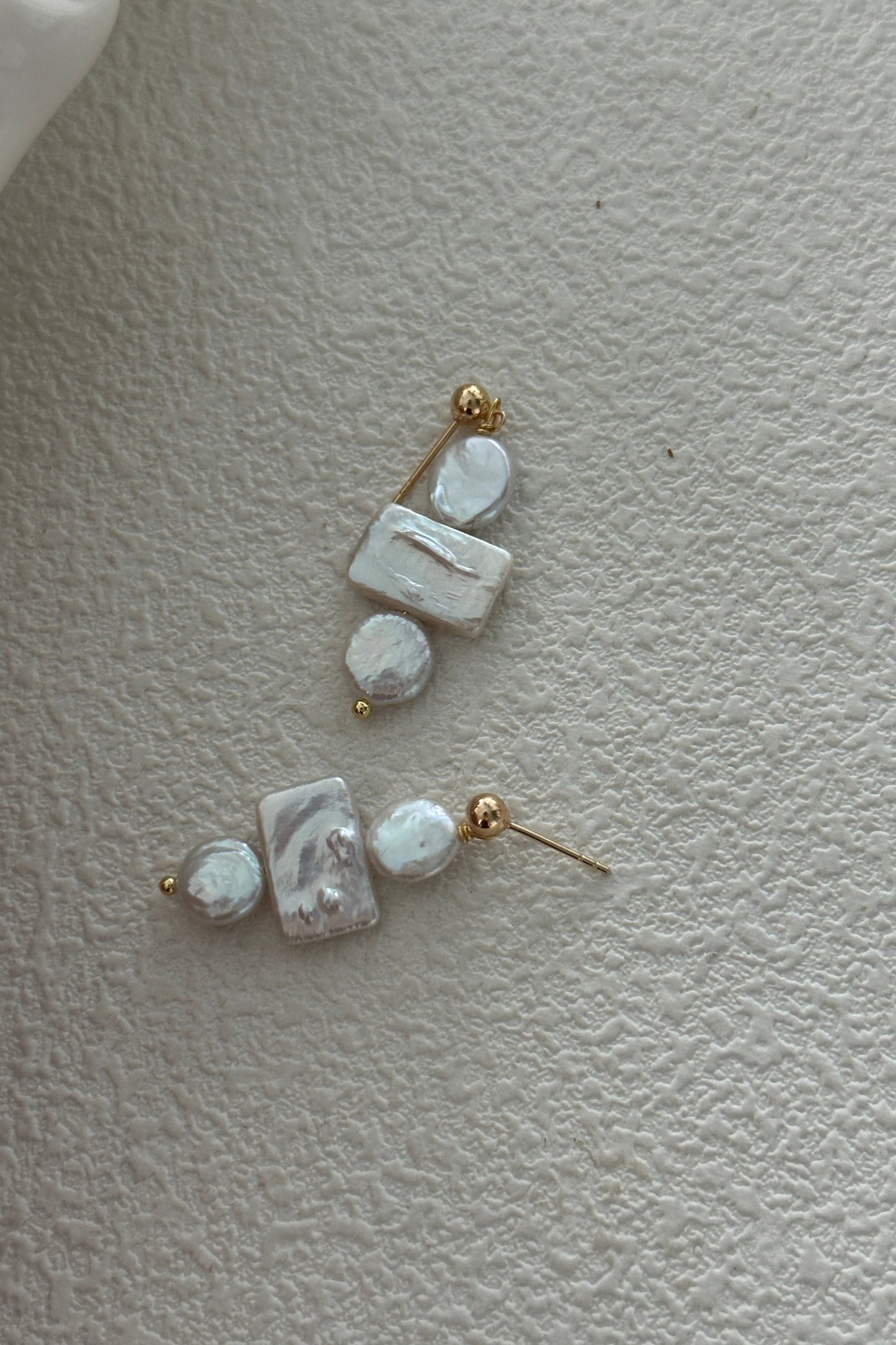 Displayed on a textured surface, the Siren Mode 18K Gold Plated 925 Silver Baroque Pearl Drop Earrings each feature a rectangular mother-of-pearl piece, a round pearl, and an irregularly shaped baroque pearl. The components are connected by small 18K gold plated beads and attached to simple gold studs.