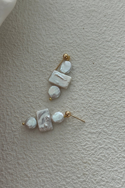 Displayed on a textured surface, the Siren Mode 18K Gold Plated 925 Silver Baroque Pearl Drop Earrings each feature a rectangular mother-of-pearl piece, a round pearl, and an irregularly shaped baroque pearl. The components are connected by small 18K gold plated beads and attached to simple gold studs.