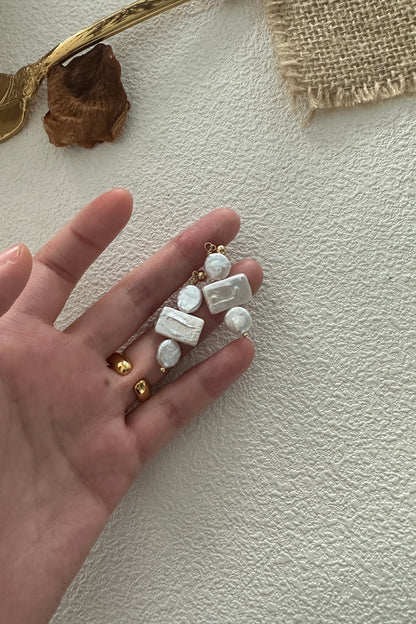 A hand holds a sophisticated pair of 18K Gold Plated 925 Silver Baroque Pearl Drop Earrings by Siren Mode, adorned with Baroque Pearls and rectangular white beads. The background includes a gold fork, a dried brown leaf, and a piece of burlap fabric on a textured white surface.