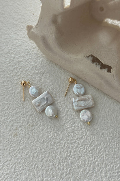 A pair of Siren Mode's 18K Gold Plated 925 Silver Baroque Pearl Drop Earrings set on a textured white surface next to a beige decorative object. Each earring features two rectangular and two round white marbled beads, arranged vertically, and attached to an 18K gold-plated stud.