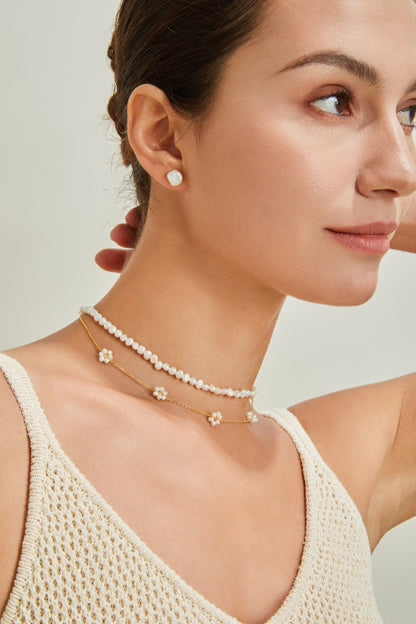 A person with tied-back hair is wearing a cream-colored knitted top. They have on two necklaces—a shorter pearl necklace and a longer 18K Gold Plated 925 Silver Flower Petal Pearl Necklace from Siren Mode. Both are 18K gold plated. They also wear a pair of small, round stud earrings.