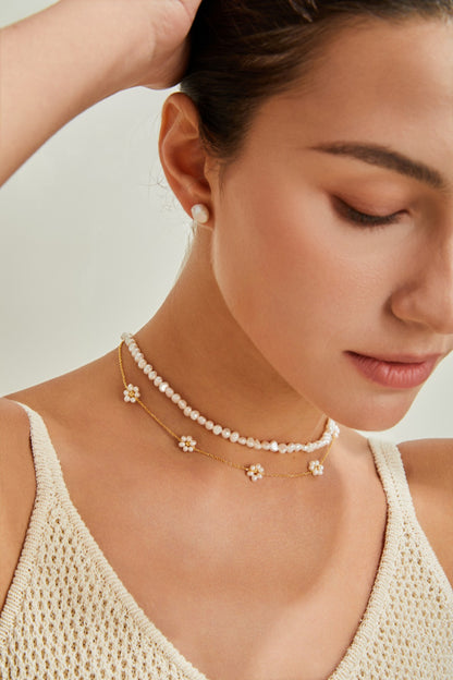 A person with their hair in a bun is wearing a delicate white-beaded pearl necklace from Siren Mode, and an 18K Gold Plated 925 Silver Flower Petal Pearl Necklace. They are also wearing a sleeveless, knitted top and a single stud earring. The person has their eyes closed and appears serene.