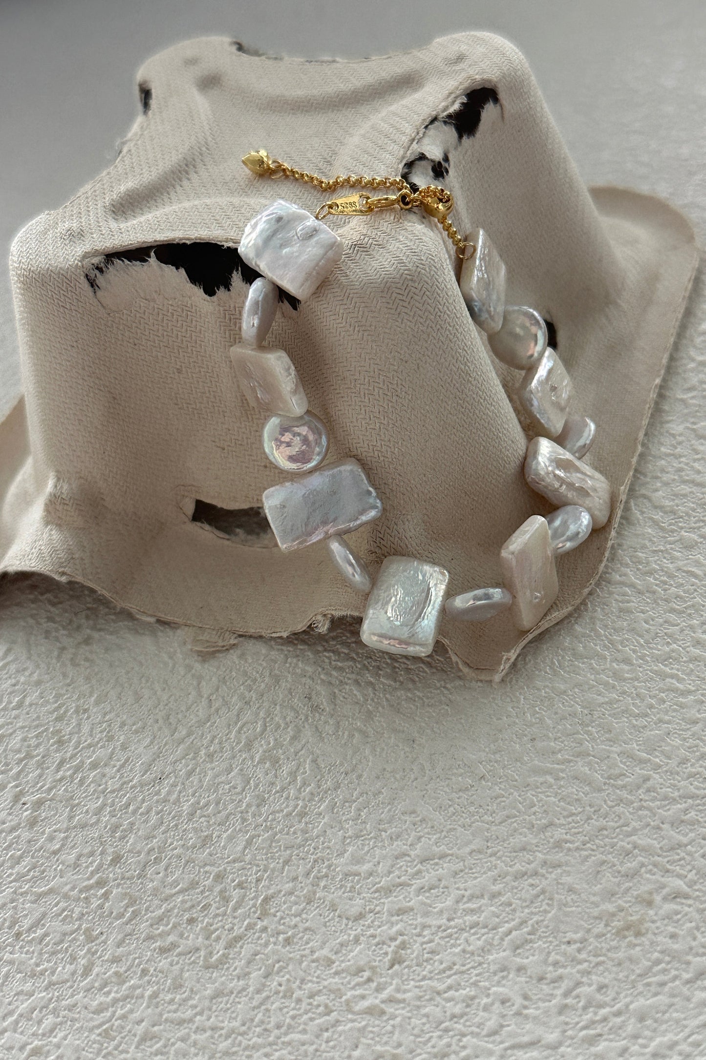 A fabric bucket hat with distressed details is displayed with a beaded necklace draped over it. The necklace, paired perfectly with the Siren Mode 18K Gold Plated 925 Silver Foil Baroque Pearl Bracelet, features irregularly shaped, pearlescent beads in varying sizes and shapes. The hat and jewelry rest on a light-colored textured surface.