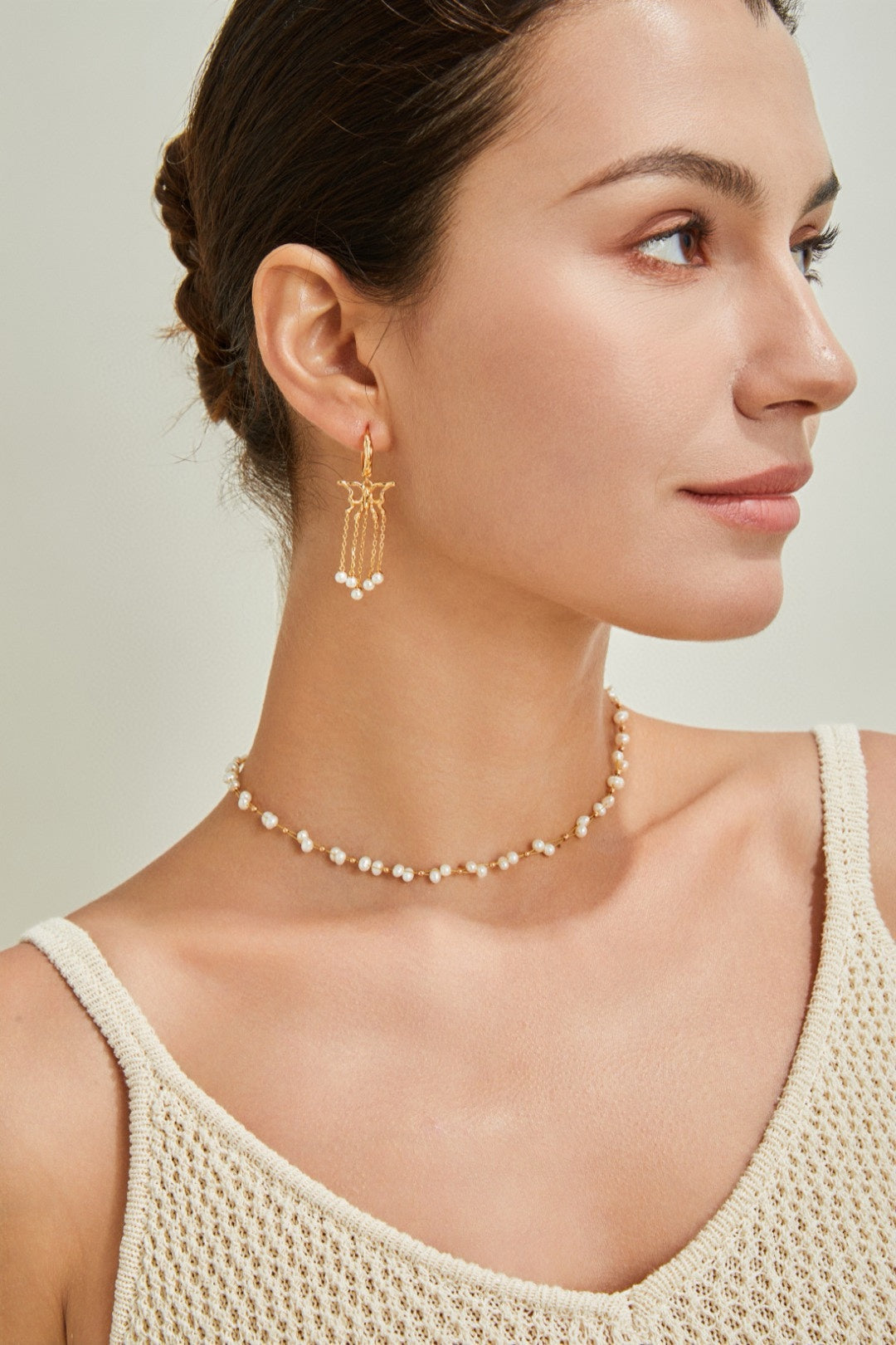 A person with dark hair pulled back into a bun is shown from the shoulders up, looking to the side. They are wearing a crochet-style beige sleeveless top, a Siren Mode 18K Gold Plated 925 Silver Handcrafted Freshwater Pearl Necklace, and 18K Gold Plated earrings with dangling pearl details. The background is neutral.