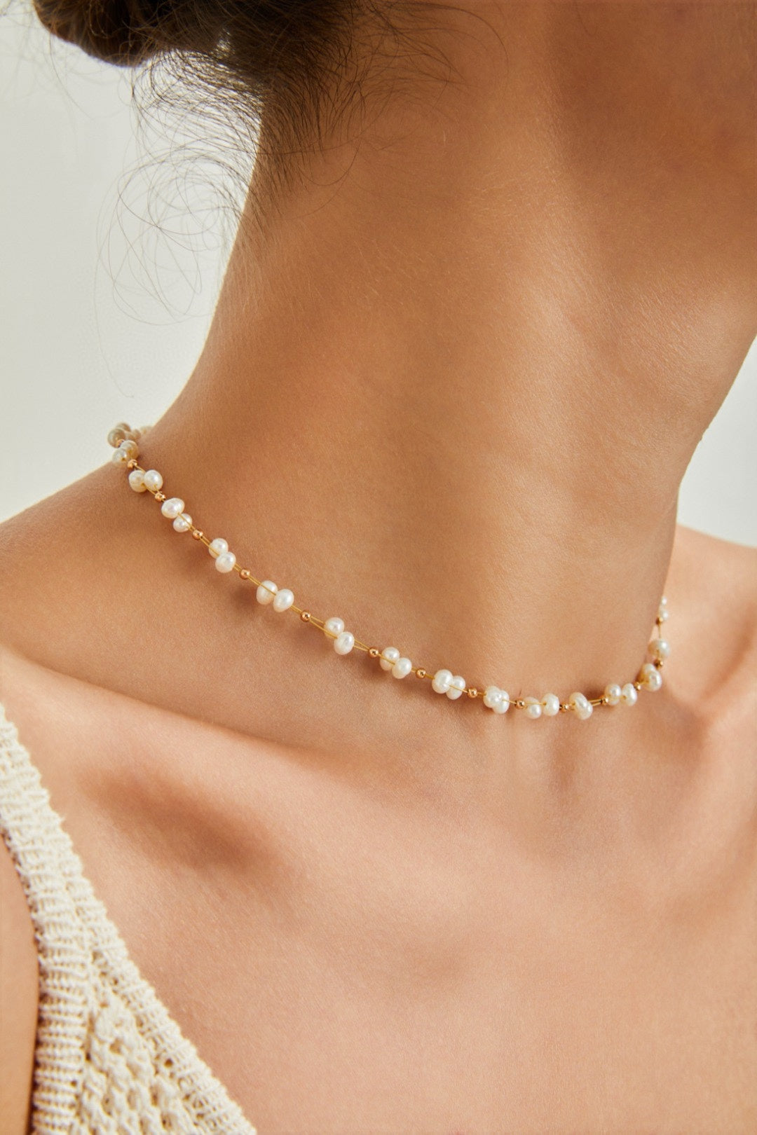 Close-up of a person's neck adorned with the exquisite Siren Mode 18K Gold Plated 925 Silver Handcrafted Freshwater Pearl Necklace, the small white pearls adding elegance. The skin is smooth, and the top of the shoulders are visible, with part of a light-colored, crocheted or knitted top. The softly out-of-focus background enhances the serene composition.