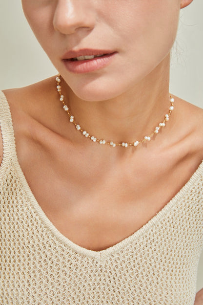 A close-up of a person wearing an 18K Gold Plated 925 Silver Handcrafted Freshwater Pearl Necklace by Siren Mode and a beige knit top. The person's face is partially visible, with the focus predominantly on the neckline and the jewelry. The neutral background keeps the attention on the necklace and the outfit.