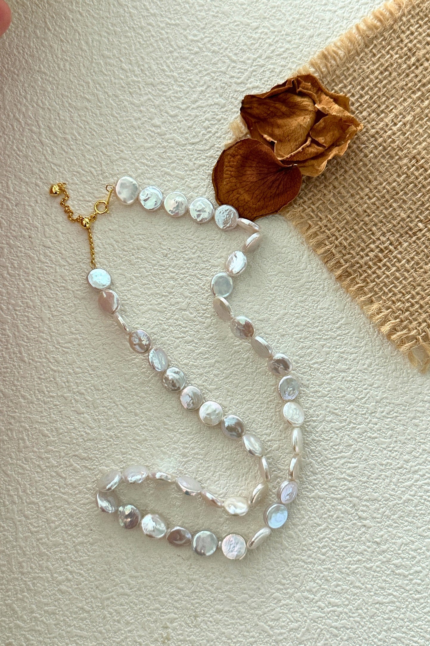 A string of iridescent white beads arranged in a loose, flowing pattern on a textured white surface. The 18K Gold Plated 925 Silver Small Round Baroque Pearl Necklace by Siren Mode features beads connected by gold plated links and a matching clasp is visible at one end. Nearby, there are some dried flower petals and a piece of burlap fabric.