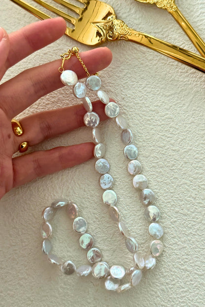 A hand is holding a Siren Mode 18K Gold Plated 925 Silver Small Round Baroque Pearl Necklace, made of round, iridescent white beads. The background features a textured surface and several ornate, gold-colored utensils. The person is wearing gold rings on their index and middle fingers, complemented by an 18K Gold Plated bracelet.