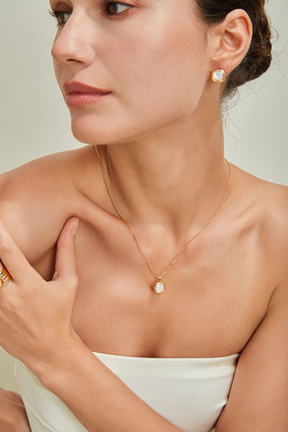 A woman with dark hair pulled back is wearing a strapless white top. She is adorned with the Siren Mode 18K Gold Plated 925 Silver Square Baroque Pearl Pendant Necklace, matching square earrings, and a gold ring on her left hand. Her skin has a smooth, glowing complexion.