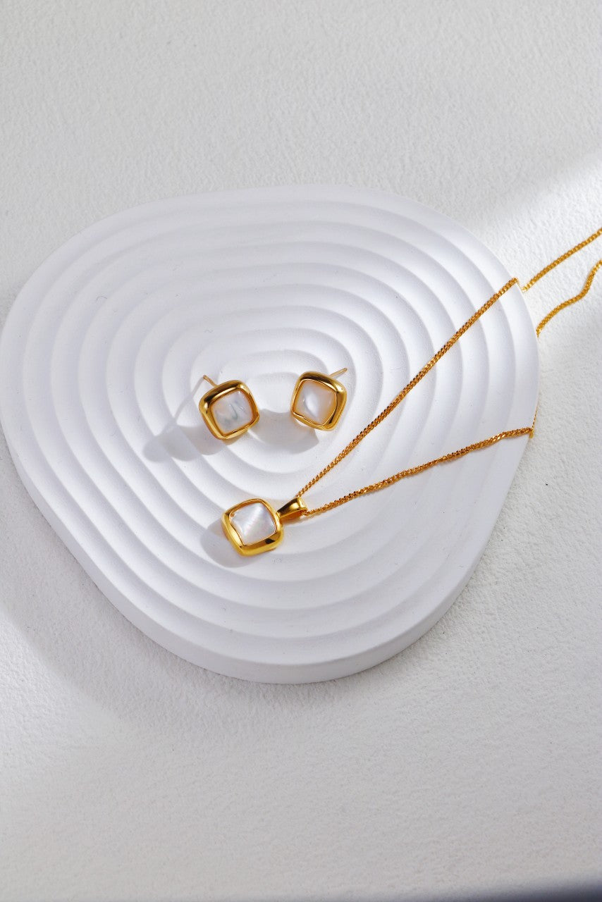 A pair of geometric 18K gold plated earrings featuring a square white stone setting and the Siren Mode 18K Gold Plated 925 Silver Square Baroque Pearl Pendant Necklace are displayed on a circular, textured white surface, with the background also being a white, textured surface.