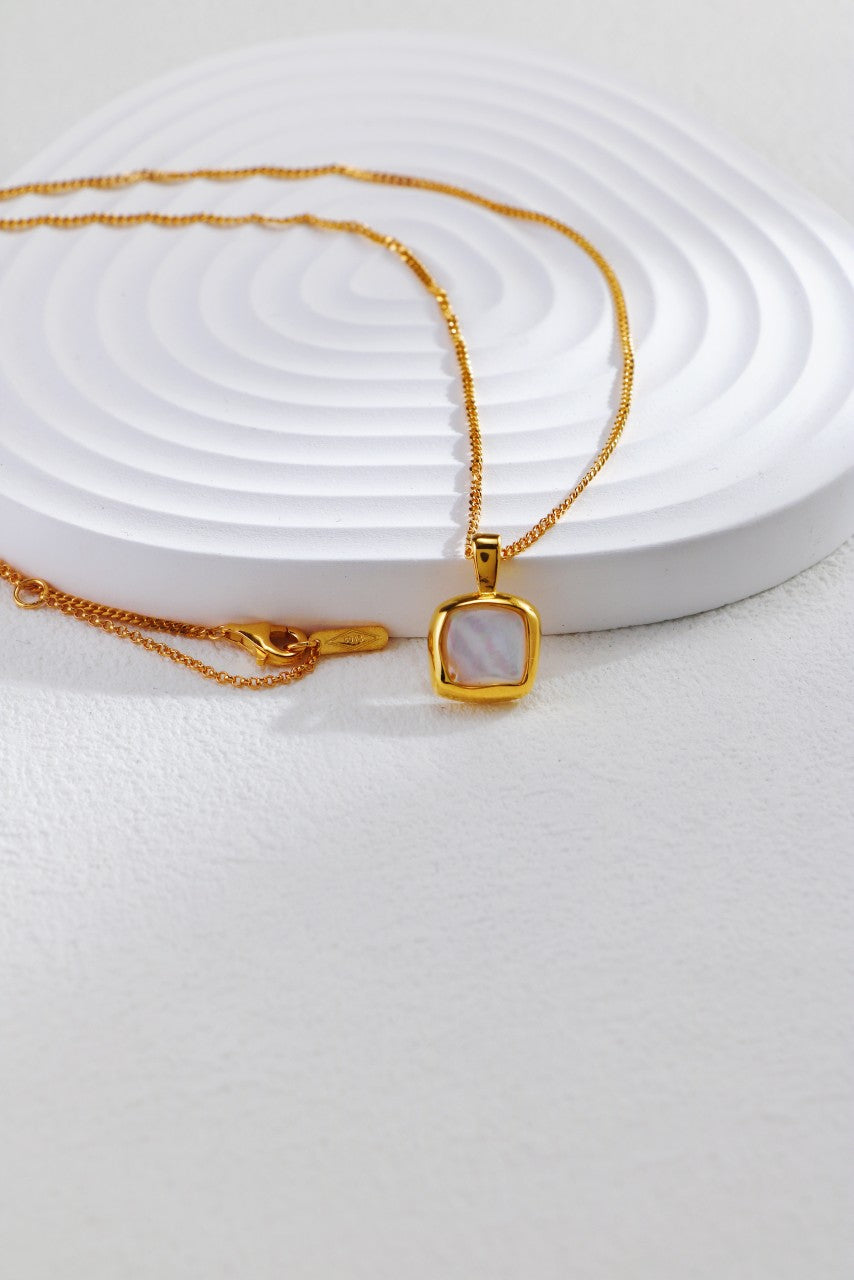 A delicate "18K Gold Plated 925 Silver Square Baroque Pearl Pendant Necklace" by Siren Mode is displayed on a white, textured, round stand with a spiral pattern. The necklace is laid out with the chain extended and the clasp visible on the left side.