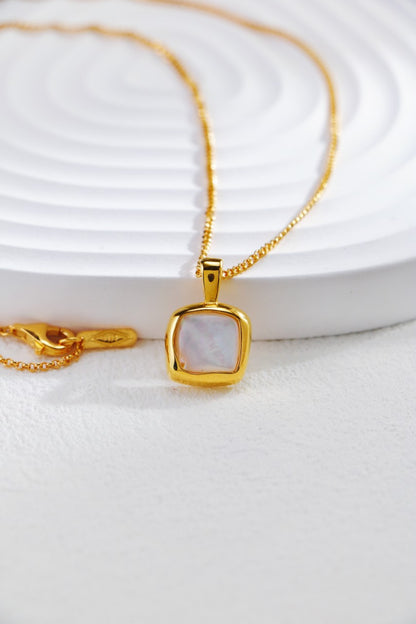 The 18K Gold Plated 925 Silver Square Baroque Pearl Pendant Necklace from Siren Mode lies delicately on a soft white textured surface. Its square-shaped pendant, featuring a luminous mother-of-pearl inlay, is prominently centered while the elegant gold chain is gracefully looped around.