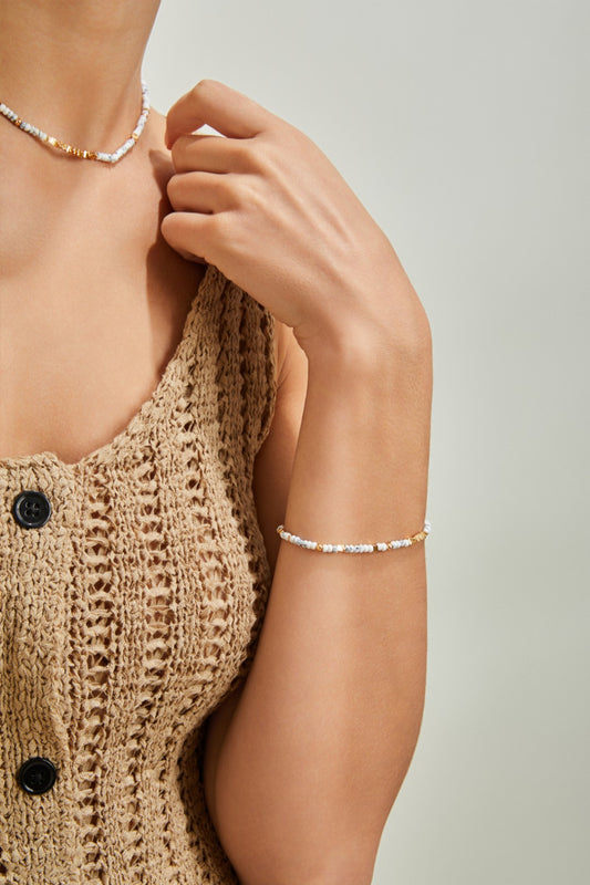 A woman wearing a beige crochet tank top with black buttons gently touches her neck with one hand. She accessorizes with a delicate 18K gold plated beaded necklace and a matching *Siren Mode* 18K Gold Plated 925 Silver White Turquoise Beaded Bracelet. The photo focuses on her upper torso and hand against a neutral background.