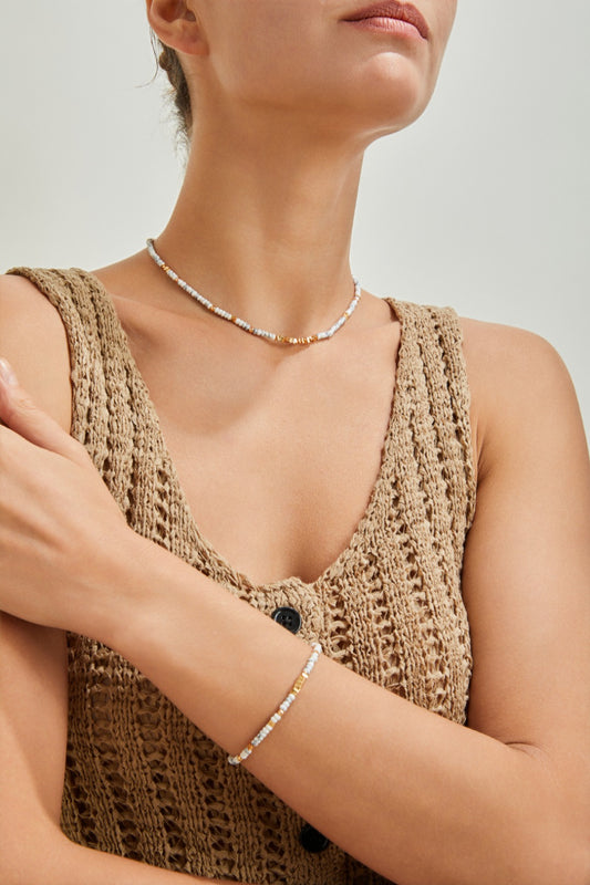 A woman showcases the Siren Mode 18K Gold Plated 925 Silver White Turquoise Beaded Necklace and its matching bracelet, both adorned with delicate white and metallic beads. She pairs them beautifully with a sleeveless, knitted beige top featuring button details. The accessories elegantly blend 925 Silver elements with the beads, all set against a plain, softly lit background.