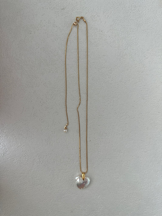 An exquisite Siren Mode Large Heart Pendant Necklace, showcasing a baroque pearl heart pendant, is displayed against a light, textured background. The chain is thin and delicate in 18K gold-plated sterling silver, featuring a subtle 14K gold clasp at one end.