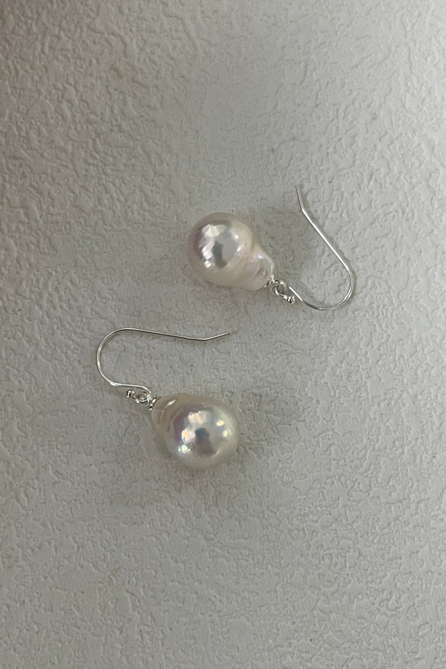 A pair of Siren Mode 925 Silver Foil Baroque Pearl Drop Earrings featuring teardrop-shaped baroque pearls with silver hooks, displayed on a textured white surface. The pearls exhibit a soft pinkish-white sheen.