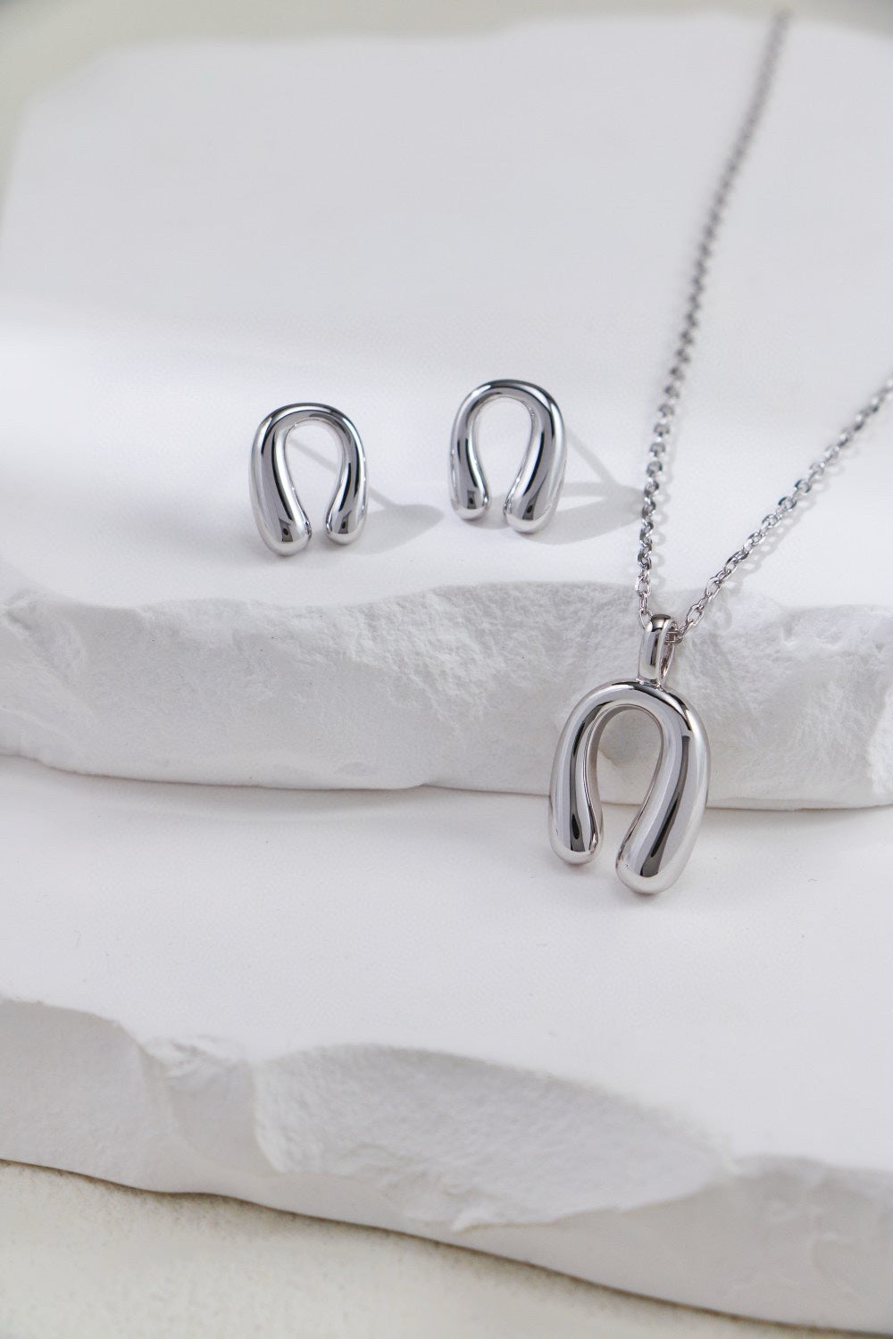 A stunning jewelry set from Siren Mode, featuring an adjustable horseshoe pendant necklace crafted in gold-plated sterling silver and a pair of minimalist horseshoe-shaped stud earrings. The elegant pieces rest on a white textured surface, with soft lighting emphasizing their shiny finish.