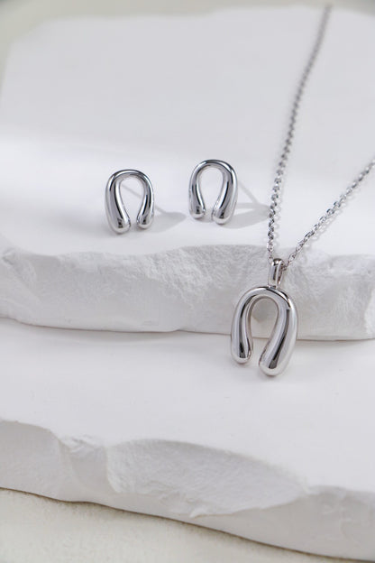 A stunning jewelry set from Siren Mode, featuring an adjustable horseshoe pendant necklace crafted in gold-plated sterling silver and a pair of minimalist horseshoe-shaped stud earrings. The elegant pieces rest on a white textured surface, with soft lighting emphasizing their shiny finish.