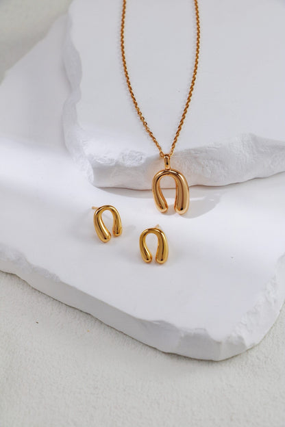 The Siren Mode Adjustable Horseshoe Pendant Necklace - Gold-Plated Sterling Silver and matching earrings are artfully arranged on layered, white, textured surfaces. The necklace features a pendant with a curved, horseshoe-like shape, and the earrings are designed to match with similar curves.