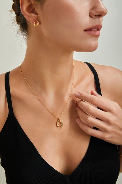 A woman wearing a sleeveless black V-neck top showcases a matching jewelry set, featuring the Siren Mode Adjustable Horseshoe Pendant Necklace in gold-plated sterling silver and small hoop earrings. She lightly touches the pendant with one hand, revealing a simple and elegant style.