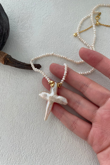 A hand holds a delicate Siren Mode Baroque Pearl Cross Necklace - Sterling Silver with 18K Gold Plating, featuring a polished, iridescent pendant. The background is a light, textured surface, and a dark, dried seed pod lies next to the hand.