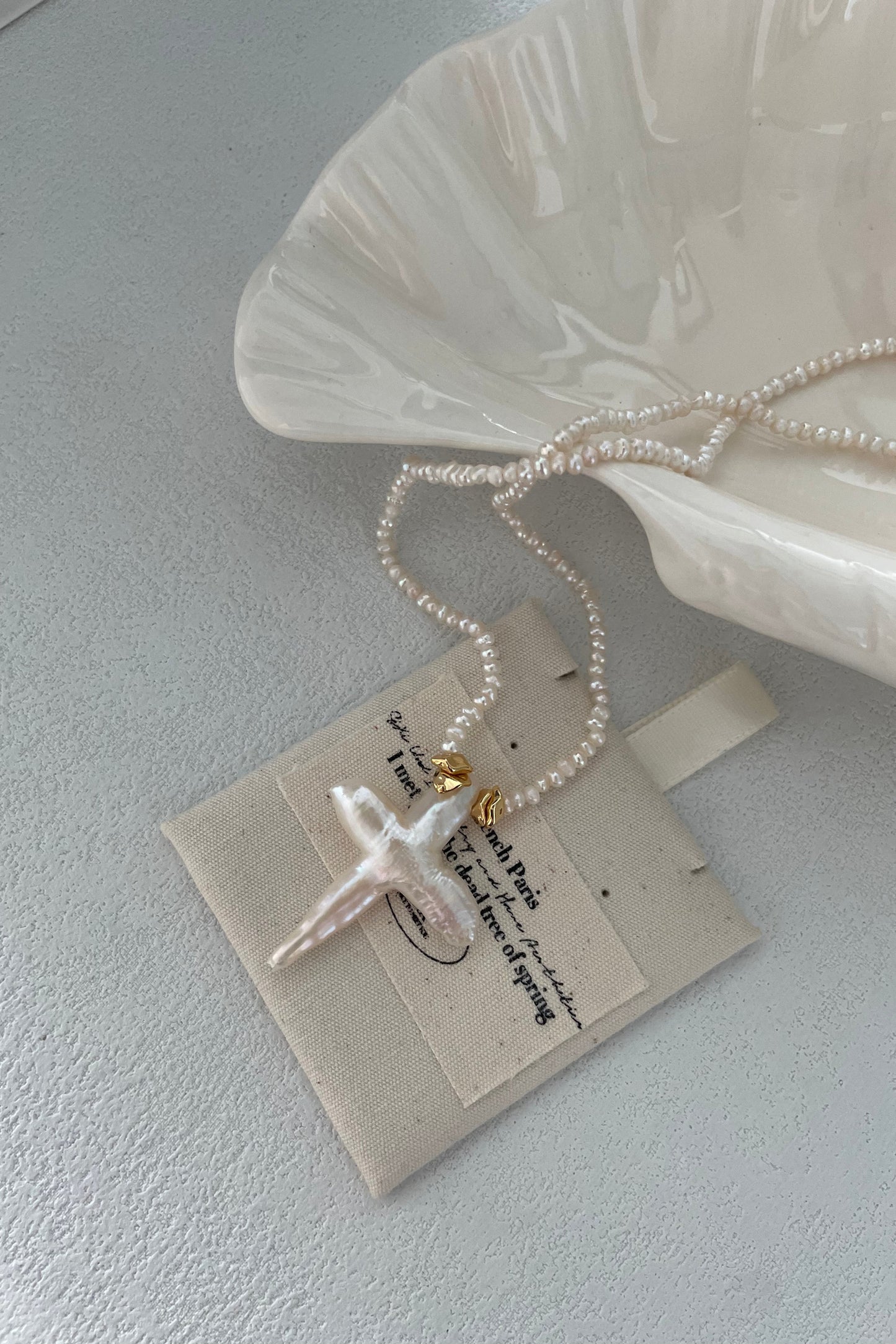 A delicate Baroque Pearl Cross Necklace with sterling silver and 18K gold plating by Siren Mode is elegantly displayed on a beige card with cursive text. The card is partially nestled inside a white, shell-like ceramic dish that boasts a subtle, glossy finish. The background features a light grey, textured surface.