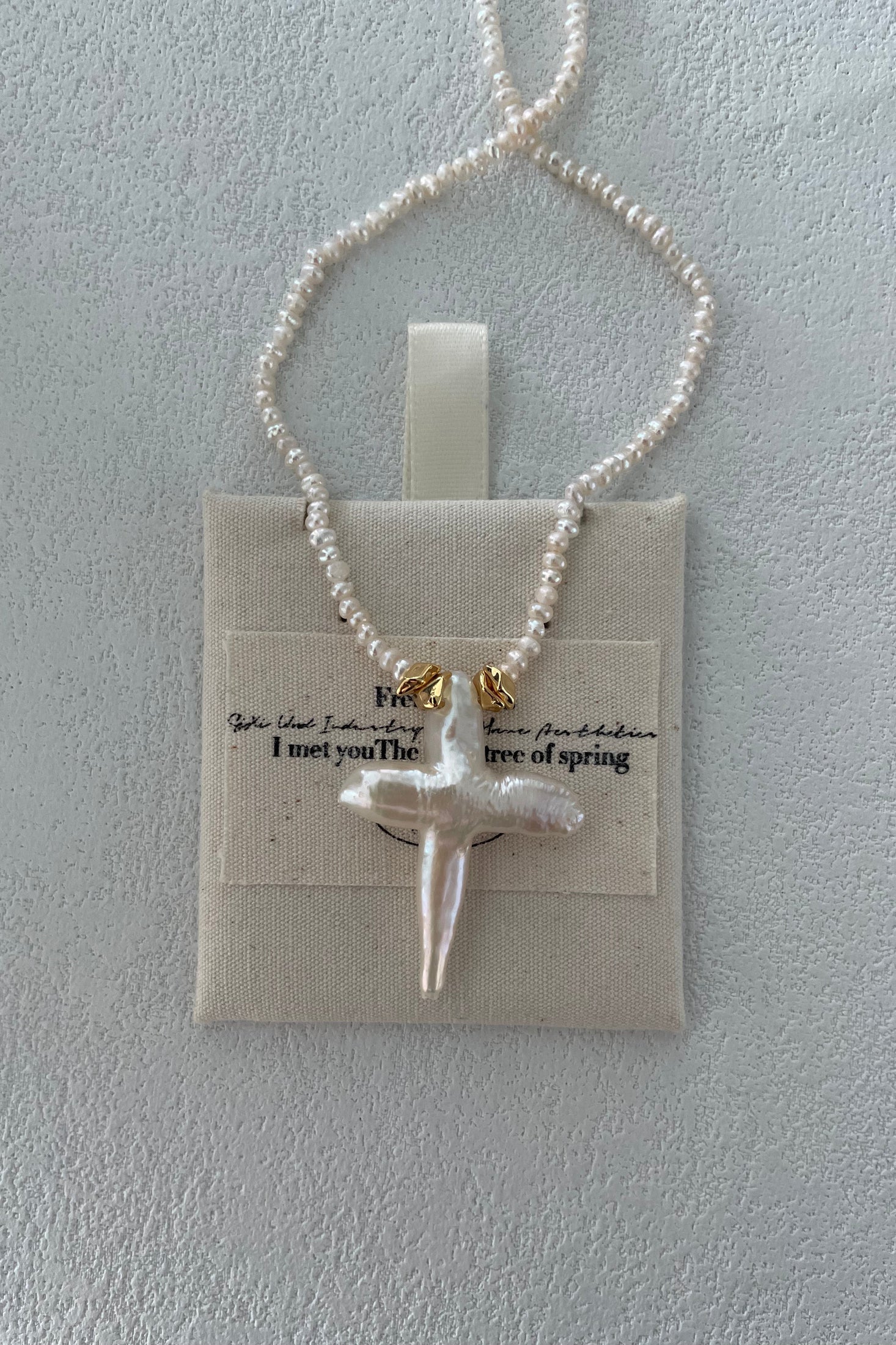 A necklace featuring small freshwater pearls and a pendant crafted from an irregularly shaped white pearl is displayed on a textured white surface. The Baroque Pearl Cross Necklace - Sterling Silver with 18K Gold Plating by Siren Mode is laid over a canvas square with the words, "I met you/The breeze of spring" partially visible.