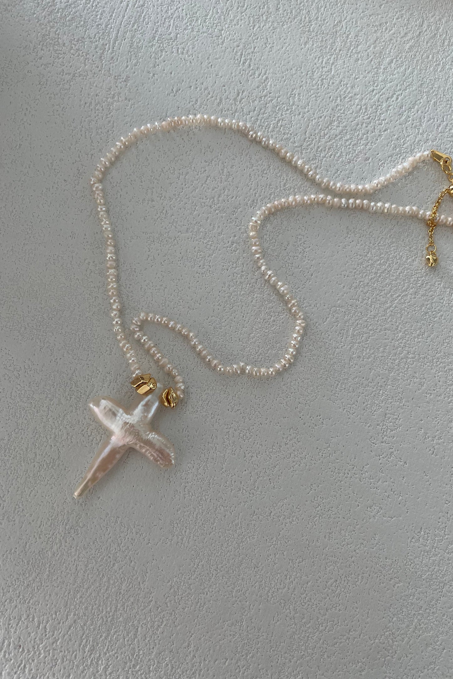 A delicate Baroque Pearl Cross Necklace by Siren Mode, featuring small freshwater pearls, is laid out on a light textured surface. The necklace has an 18K gold-plated sterling silver clasp and a pendant shaped like a cross, crafted from an irregular, translucent material.