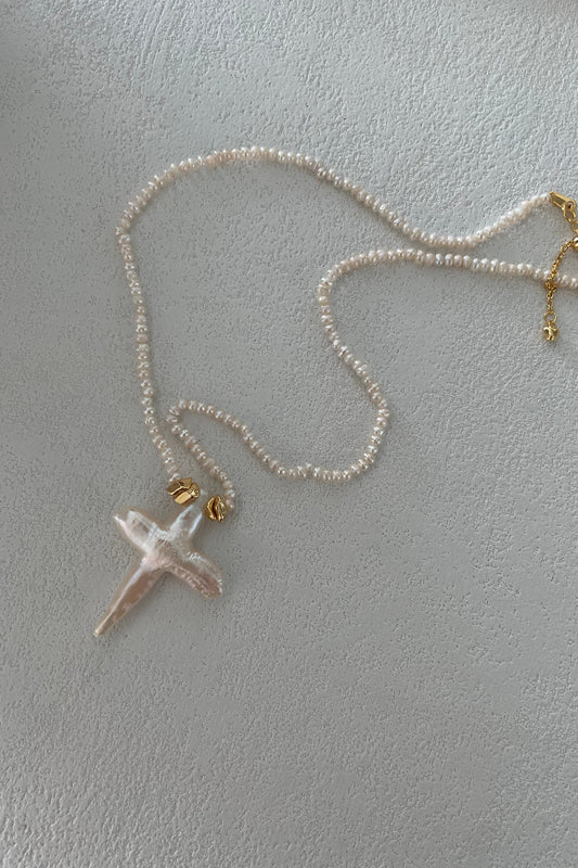 A delicate Baroque Pearl Cross Necklace by Siren Mode, featuring small freshwater pearls, is laid out on a light textured surface. The necklace has an 18K gold-plated sterling silver clasp and a pendant shaped like a cross, crafted from an irregular, translucent material.