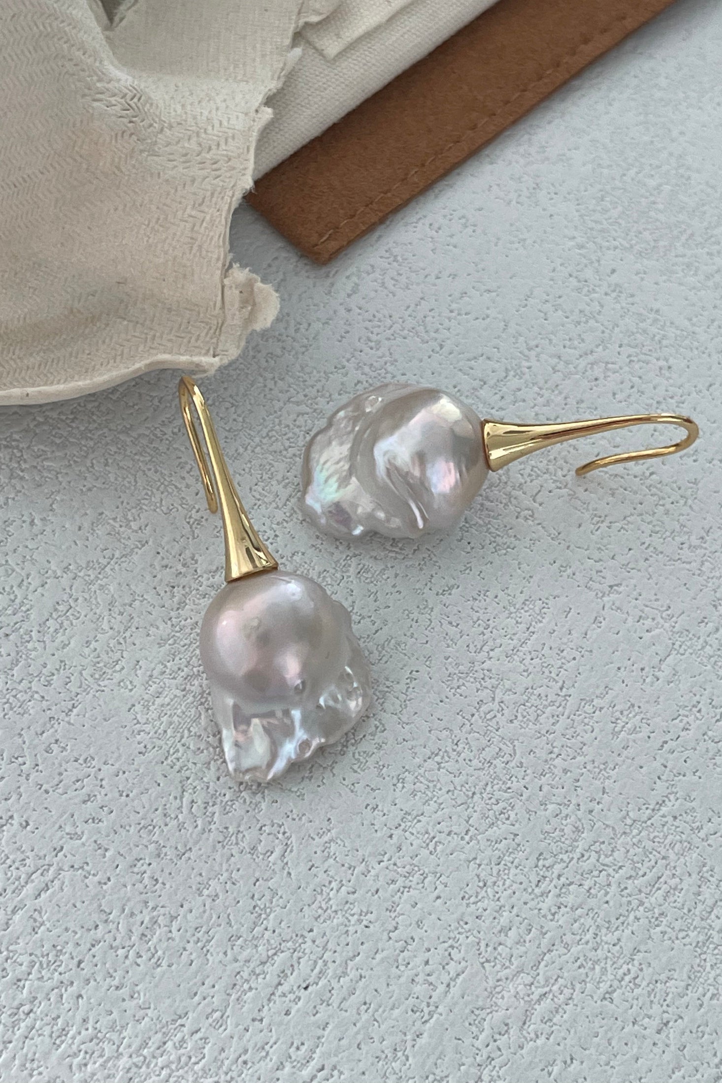 A pair of Siren Mode Baroque Pearl Drop Earrings - 18k Gold Plated Silver, featuring irregularly shaped, luminescent pearls, dangles elegantly from sleek gold-plated hooks on a light gray surface with off-white fabric and brown leather in the background.