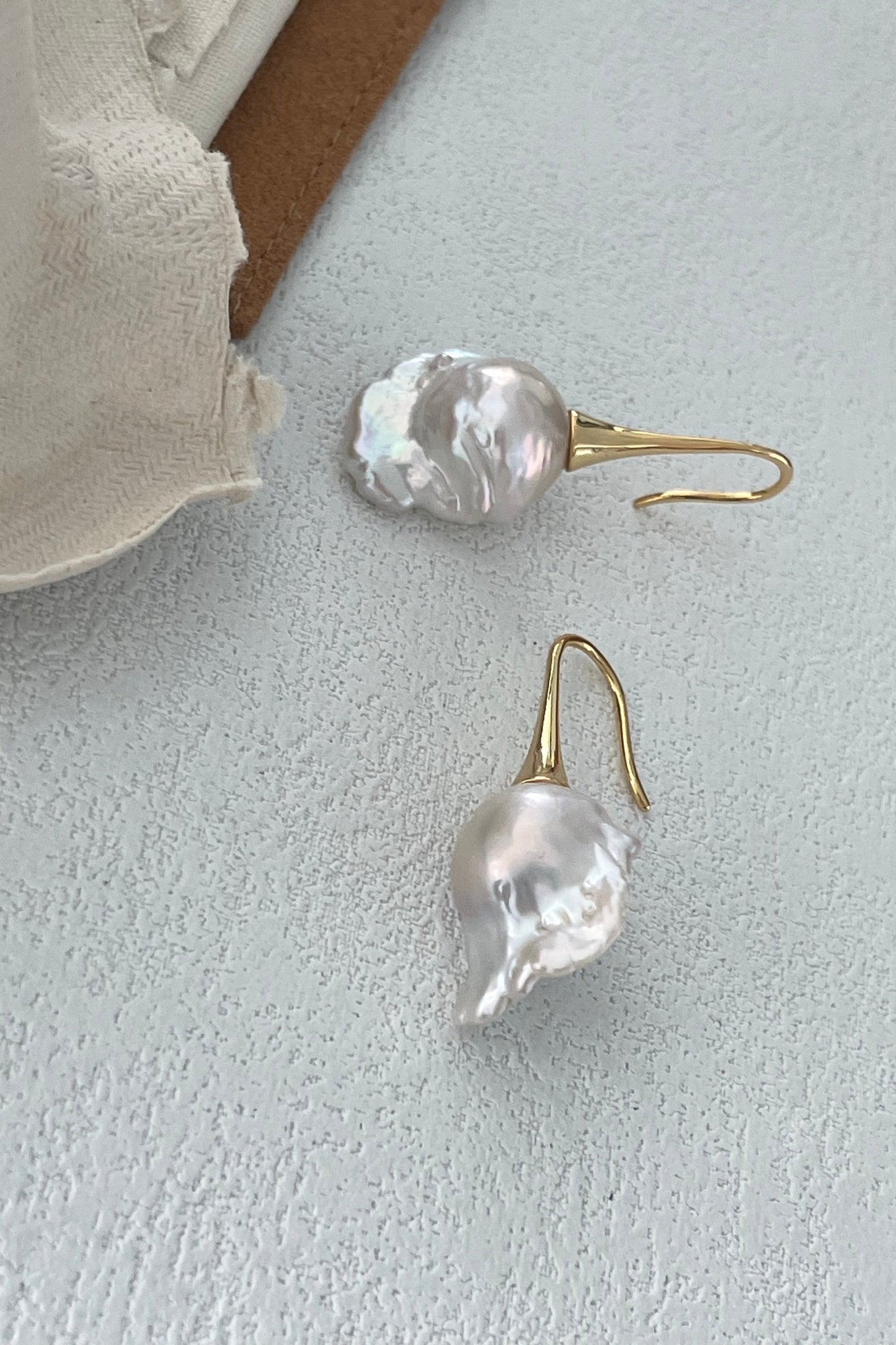 The Siren Mode Baroque Pearl Drop Earrings - 18k Gold Plated Silver are artfully displayed on a light gray textured surface. These exquisite earrings, featuring irregularly shaped baroque pearls set in gold plated silver, are positioned next to a piece of off-white fabric that is partially visible in the upper left corner.