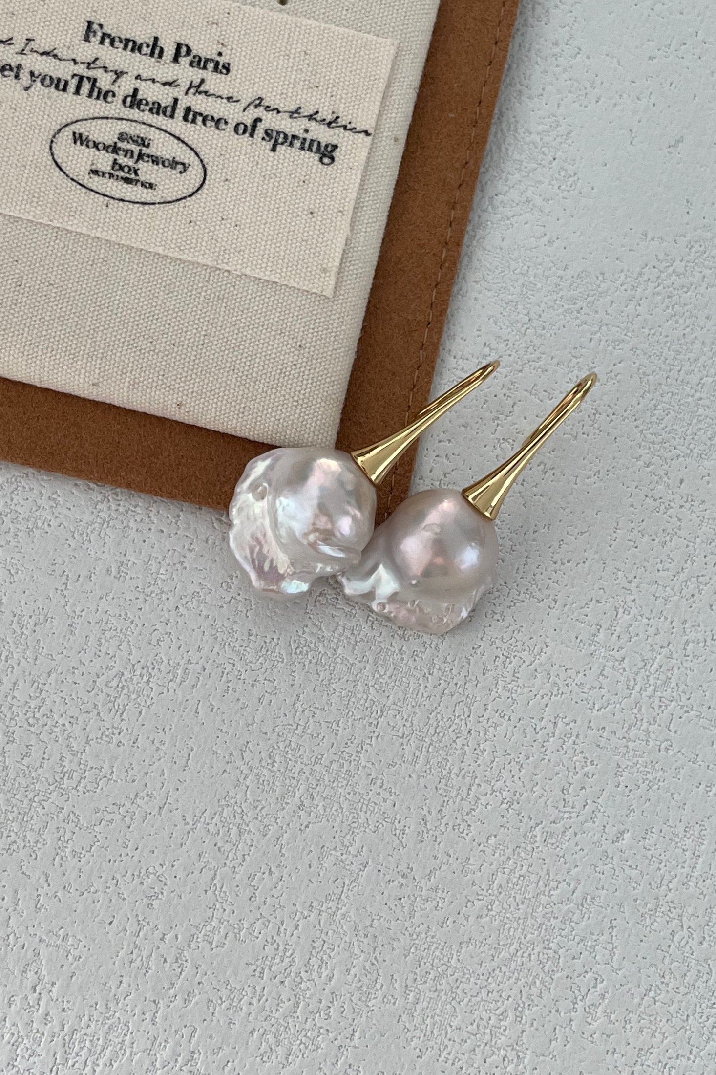 The Baroque Pearl Drop Earrings - 18k Gold Plated Silver by Siren Mode rest elegantly on a textured white surface near the edge of a brown and white embroidered fabric label with partially visible text.