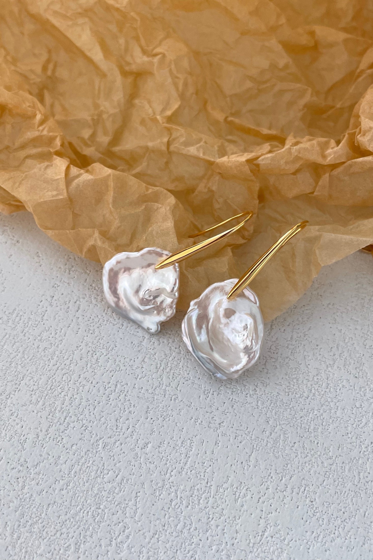 The Siren Mode Baroque Pearl Drop Earrings, featuring sterling silver hooks with 18K gold plating, are displayed on crumpled brown paper set against a light gray textured surface. The pearls showcase their natural, organic form, enhancing the earrings' unique and elegant appearance.