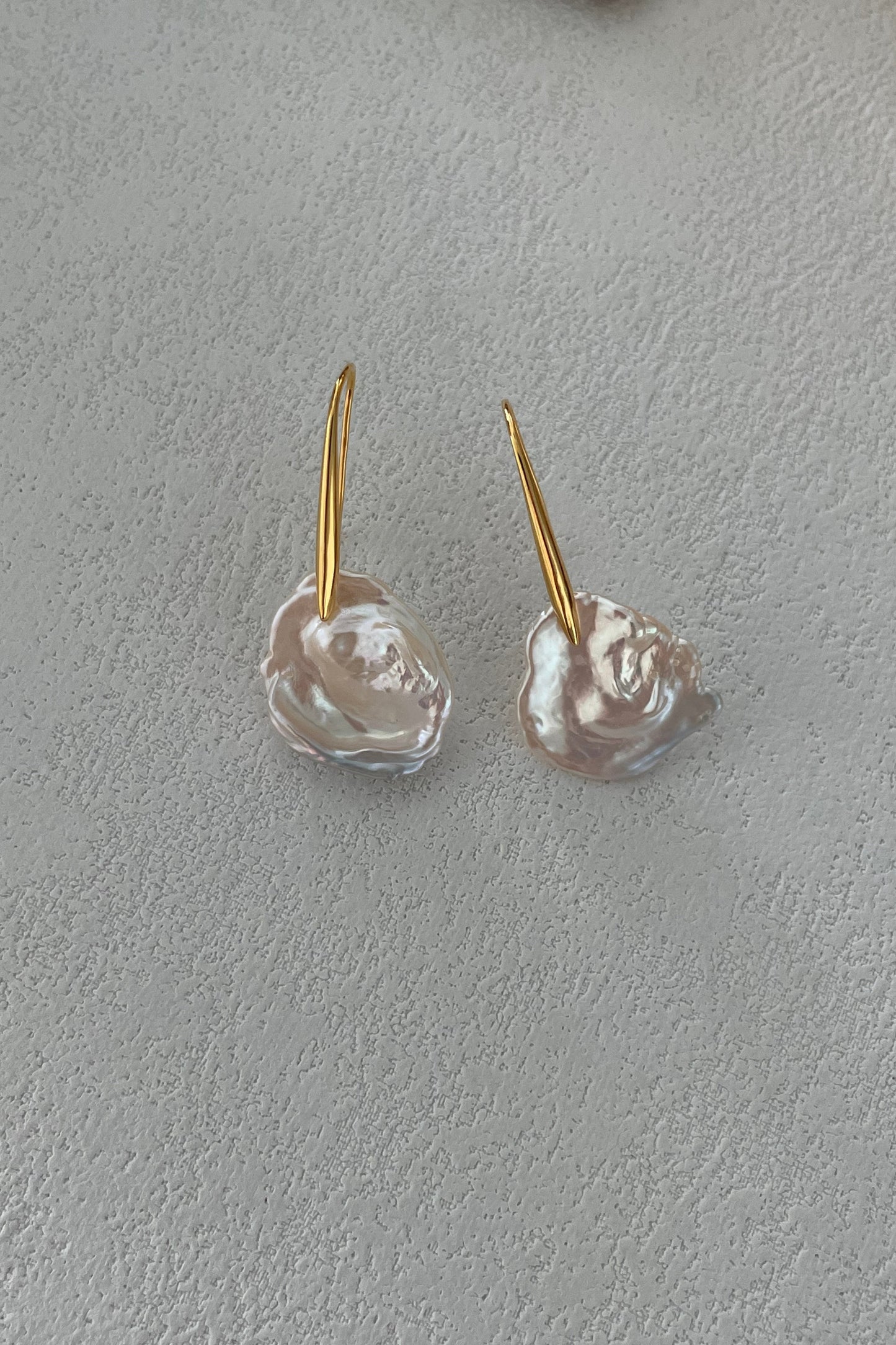 A pair of Baroque Pearl Drop Earrings from Siren Mode, featuring irregularly shaped, textured white pendants on sterling silver hooks with 18K gold plating, hang elegantly on a light gray textured surface.