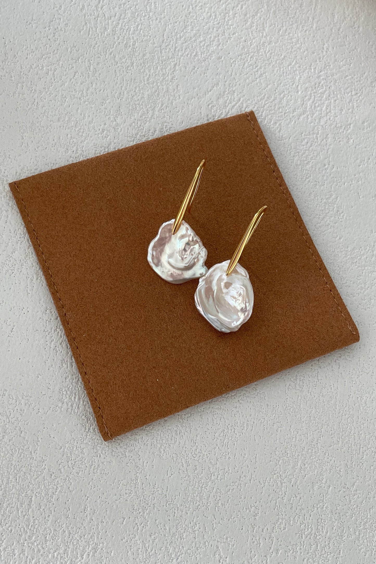 A pair of Siren Mode's Baroque Pearl Drop Earrings - Sterling Silver with 18K Gold Plating, featuring white, irregularly shaped floral-like attachments hanging from gold hooks, is displayed on a brown square piece of material against a light, textured background.