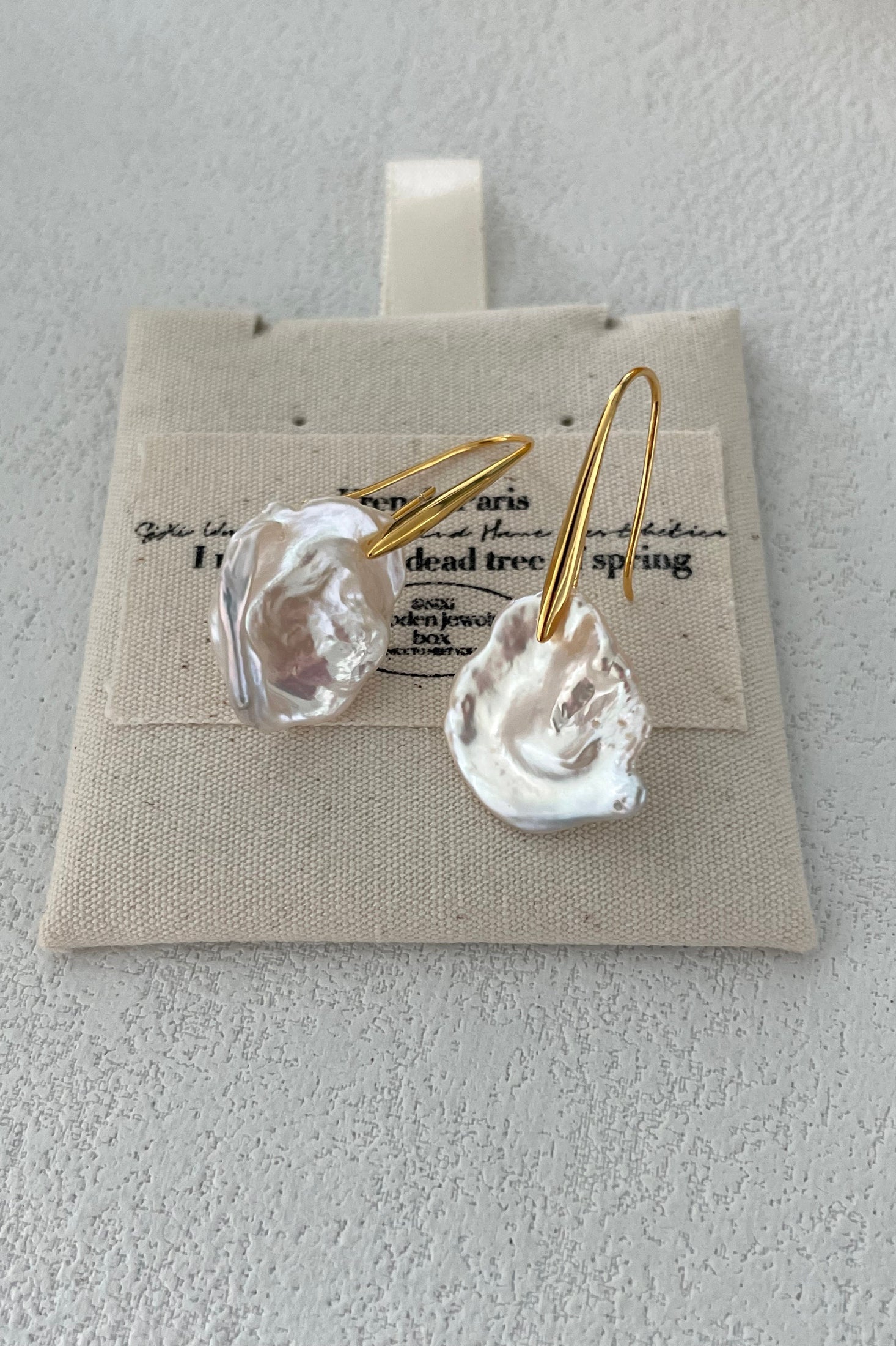 A close-up of Siren Mode's elegant Baroque Pearl Drop Earrings, featuring sterling silver hooks with 18K gold plating and irregularly shaped, glossy white natural pearls, displayed on a beige fabric with faint printed text. The background is a textured light gray surface.