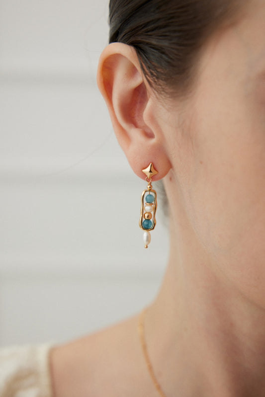 Close-up of a woman's ear wearing the Celestial Blue and Pearl Drop Earrings by Siren Mode. These stunning earrings feature a vintage gold star-shaped stud and a rectangular section adorned with celestial blue stones and lustrous pearls, perfect for a refined daytime look. The background is softly blurred.