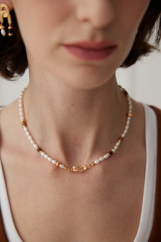 Close-up image of a person wearing the Classic Pearl and Tiger's Eye Necklace in Vintage Gold by Siren Mode. The delicate necklace features natural pearls, tiger's eye, and gold beads, centered around an ornate gold clasp. The individual is also wearing a white top layered with a brown garment, and a 925 silver earring is partially visible.