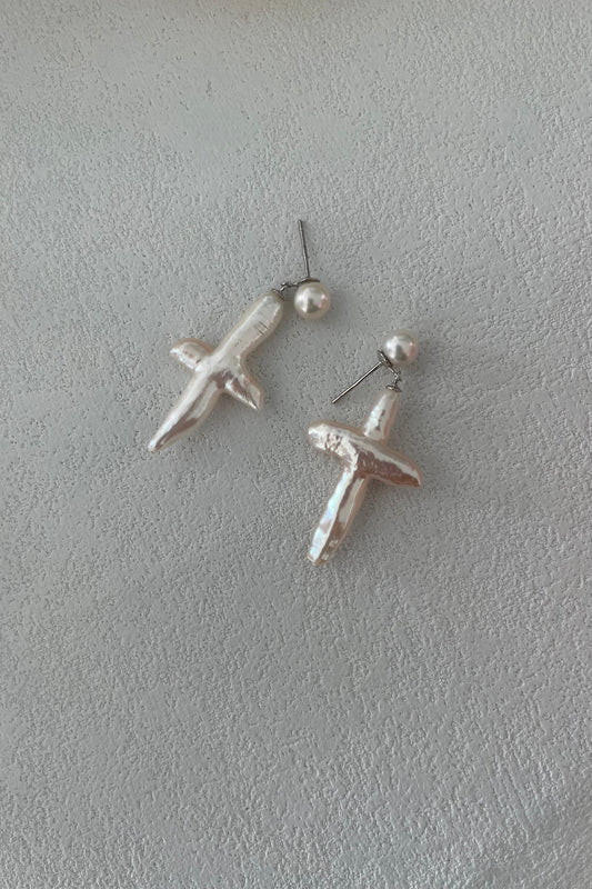 The Cross-Shaped Baroque Pearl Earrings by Siren Mode, featuring dangling pendants set on sterling silver posts, are displayed on a pale textured surface.