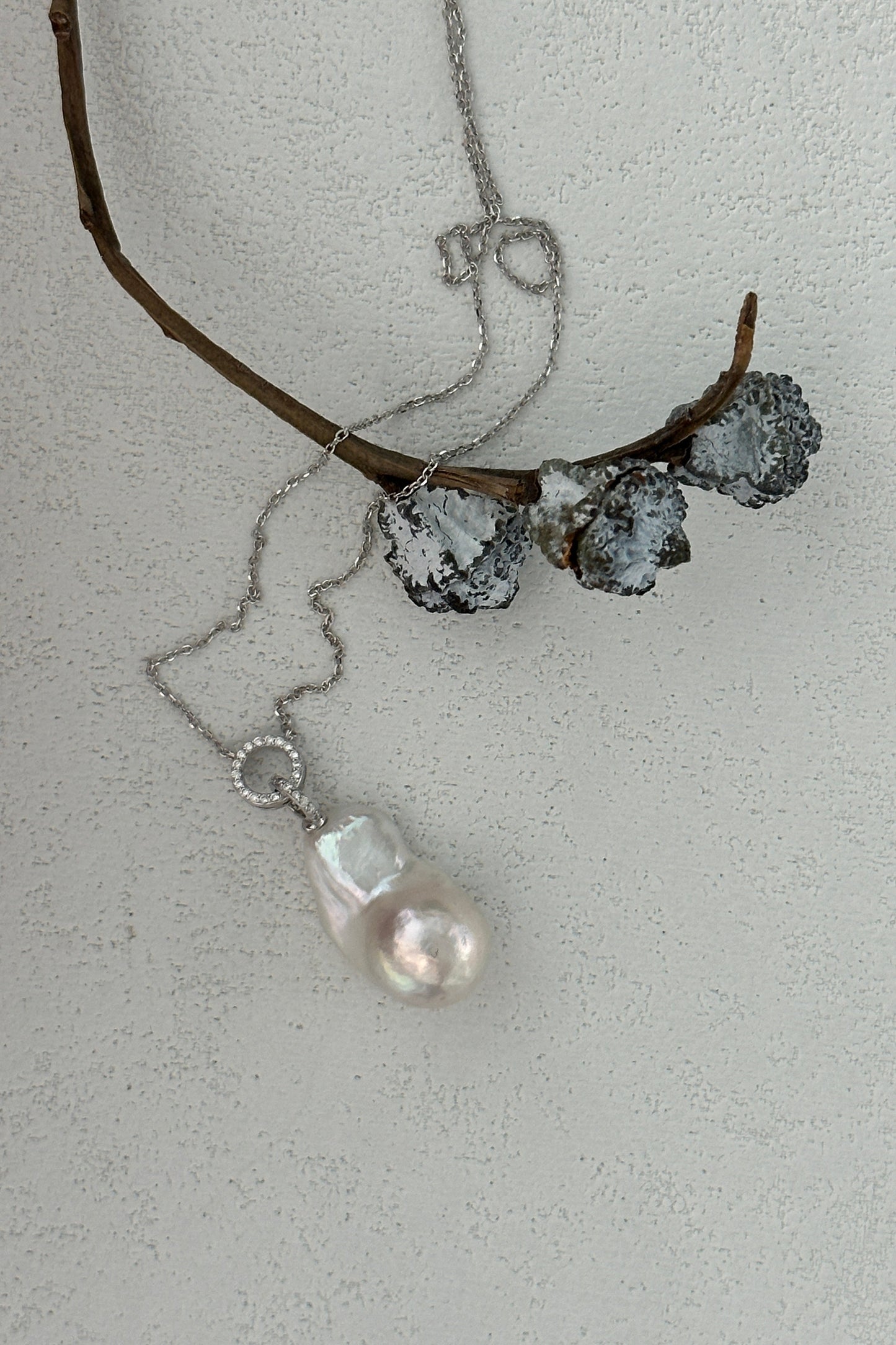 The Siren Mode Double Circle Crystal Baroque Pearl Necklace - Sterling Silver, featuring a delicate necklace with a large baroque pearl pendant accented by a small crystal, is laid on a textured, light-colored surface. The sterling silver chain gently drapes near a twig adorned with three frosted, silver-toned cones.