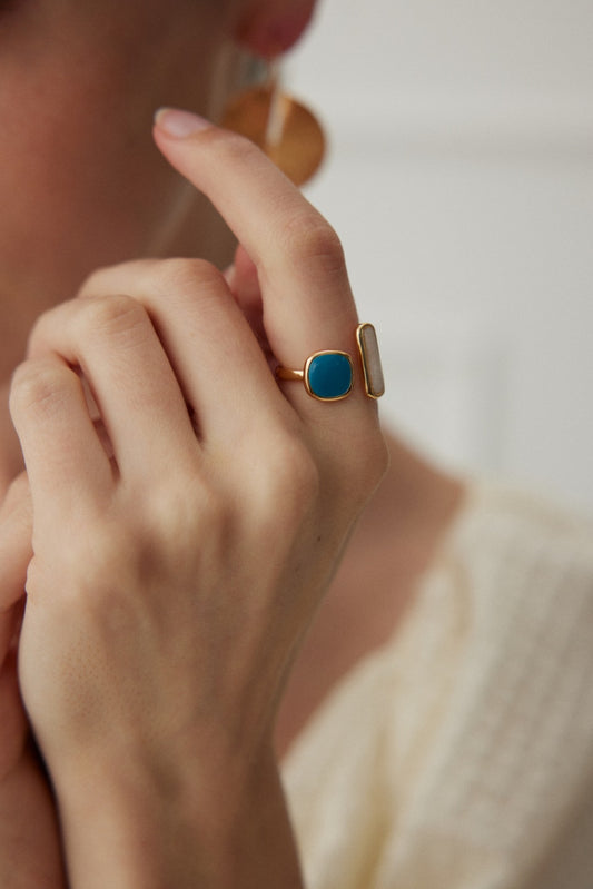 Close-up of a person touching their face while wearing the Siren Mode Dual Charm Adjustable Ring in vintage gold with an enamel finish, featuring a large blue stone and a vertical rectangular stone. The person is also wearing a large circular earring, and their hand is positioned near their ear, highlighting the jewelry.