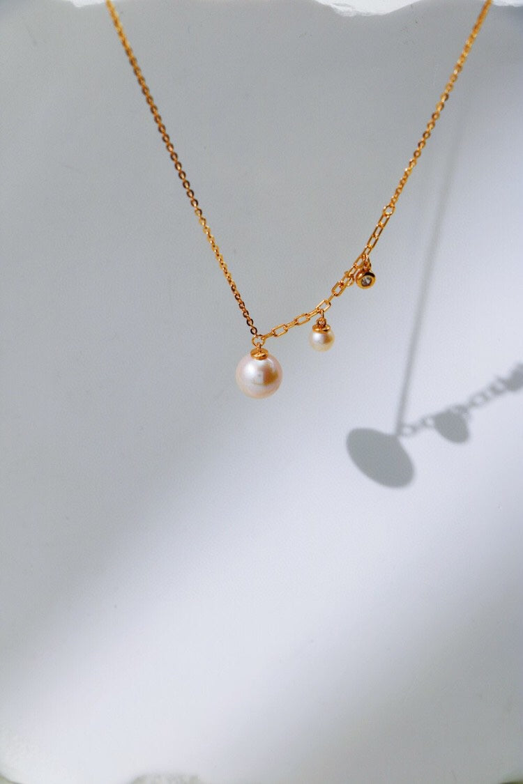 The Elegant Freshwater Pearl Necklace from Siren Mode, crafted in sterling silver, is displayed against a white backdrop. The necklace features two hanging freshwater pearls of different sizes and a small gold charm. The shadows of the pearls and chain are softly cast on the surface.
