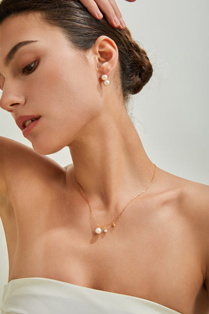 A person with dark hair styled in a low bun is shown in a close-up, profile view. They are wearing pearl earrings and the Elegant Freshwater Pearl Necklace by Siren Mode, crafted from sterling silver. The individual is wearing a strapless top and their right hand, adorned with rings of vintage gold finish, is raised, touching their hair.