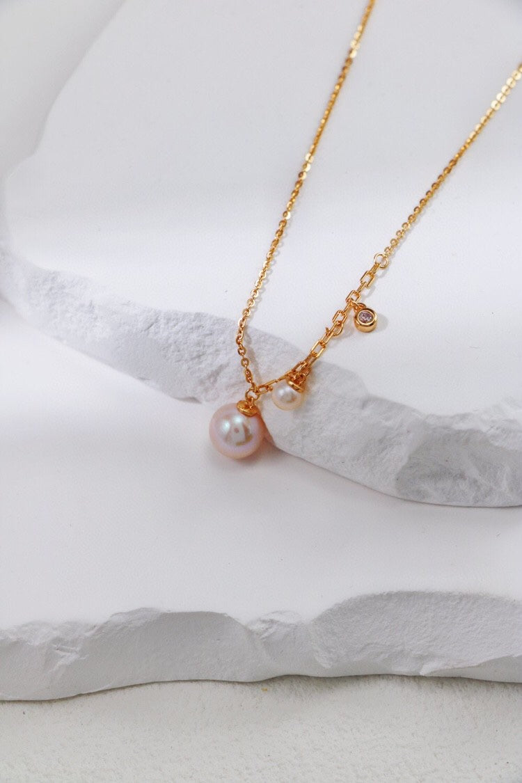 The Elegant Freshwater Pearl Necklace from Siren Mode, crafted in sterling silver and featuring two small freshwater pearls with a tiny gemstone pendant, is elegantly draped over textured white stone slabs in a minimalist setting.