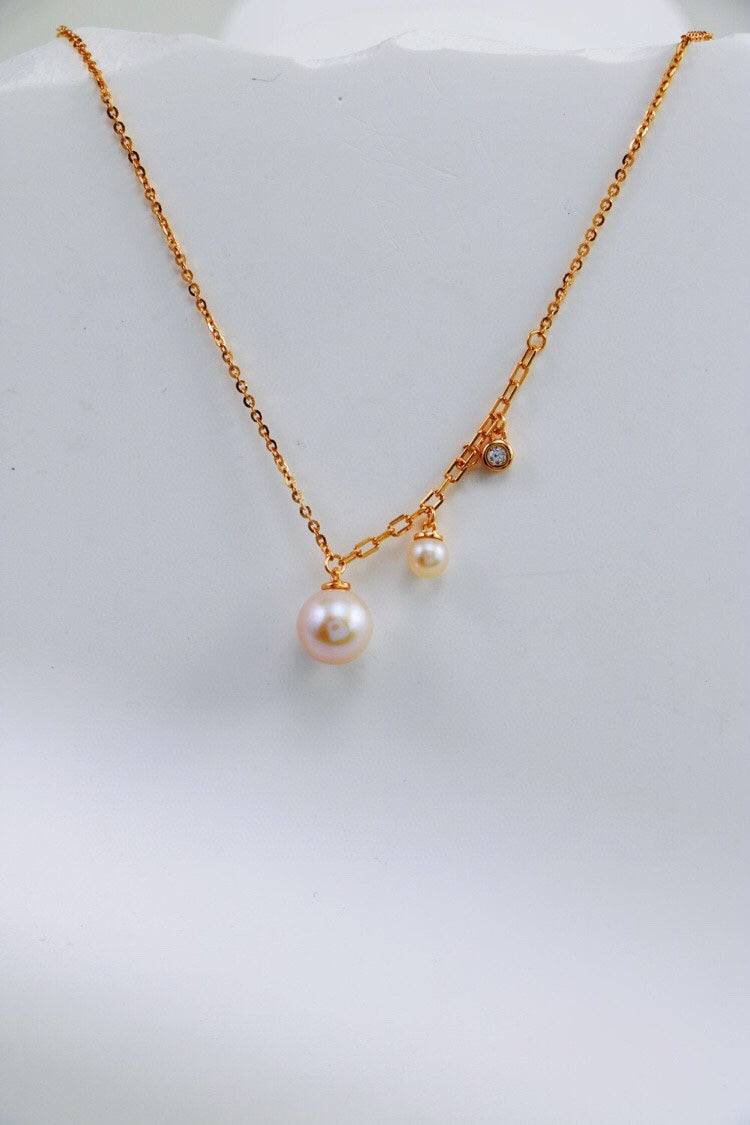 Siren Mode's Elegant Freshwater Pearl Necklace in Sterling Silver features a minimalist design with two dangling freshwater pearls of different sizes and a small, glittering diamond accent. The smooth, white surface backdrop highlights the elegance and simplicity of this exquisite jewelry piece.