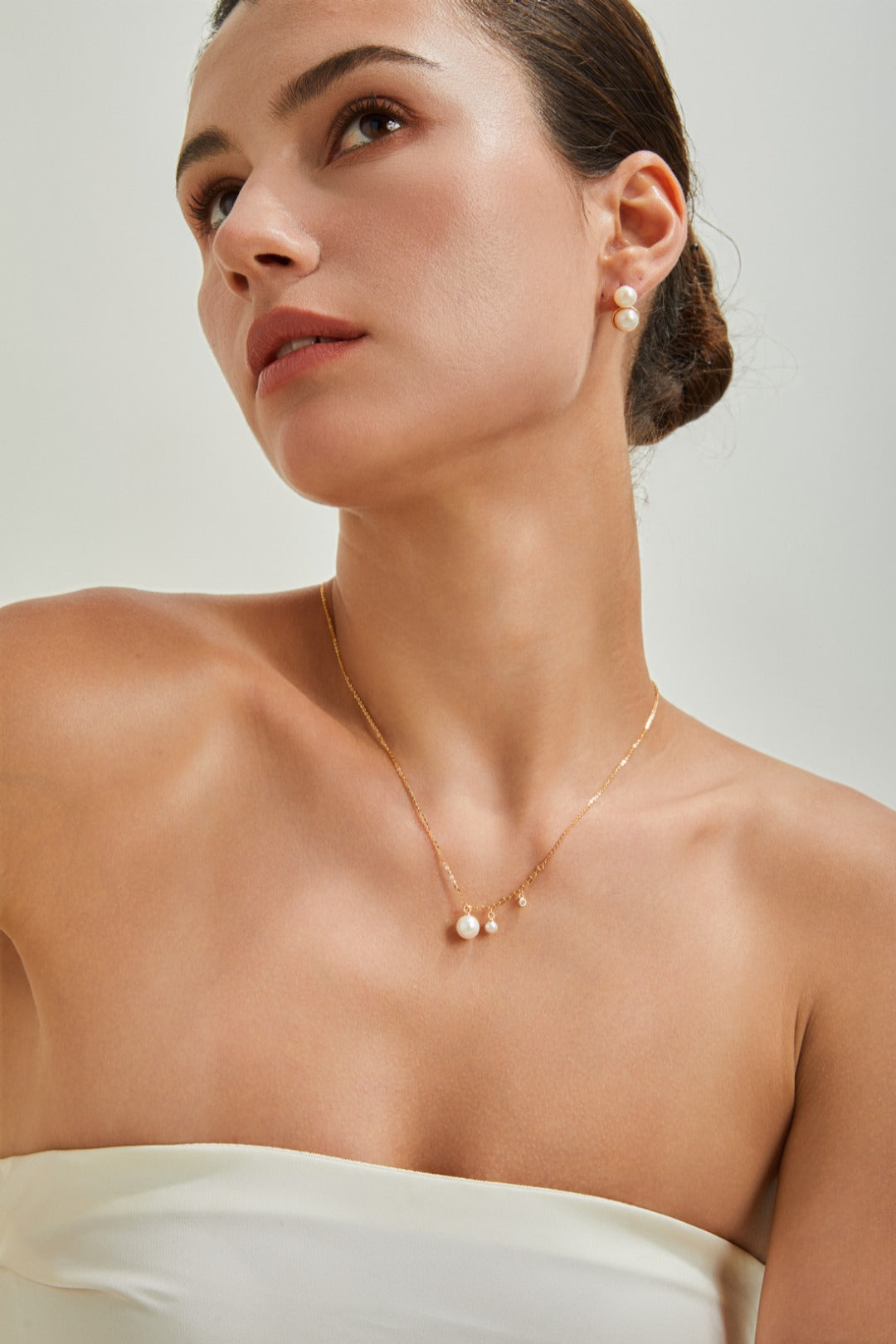 A woman with her hair styled in an updo is wearing pearl drop earrings and a delicate Siren Mode Elegant Freshwater Pearl Necklace - Sterling Silver. She has a calm, contemplative expression and is dressed in a strapless white top. The background is a simple, light neutral shade, emphasizing the sterling silver accents.