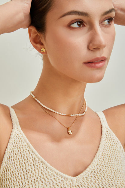 A woman with her arms raised behind her head, wearing a sleeveless, light-colored knit top. She has small, circular gold earrings and two necklaces: a white beaded choker and an adjustable length necklace called the "Elegant Gold-Plated Sterling Silver Necklace with Teardrop Pendant" by Siren Mode. She has a calm expression against a neutral background.