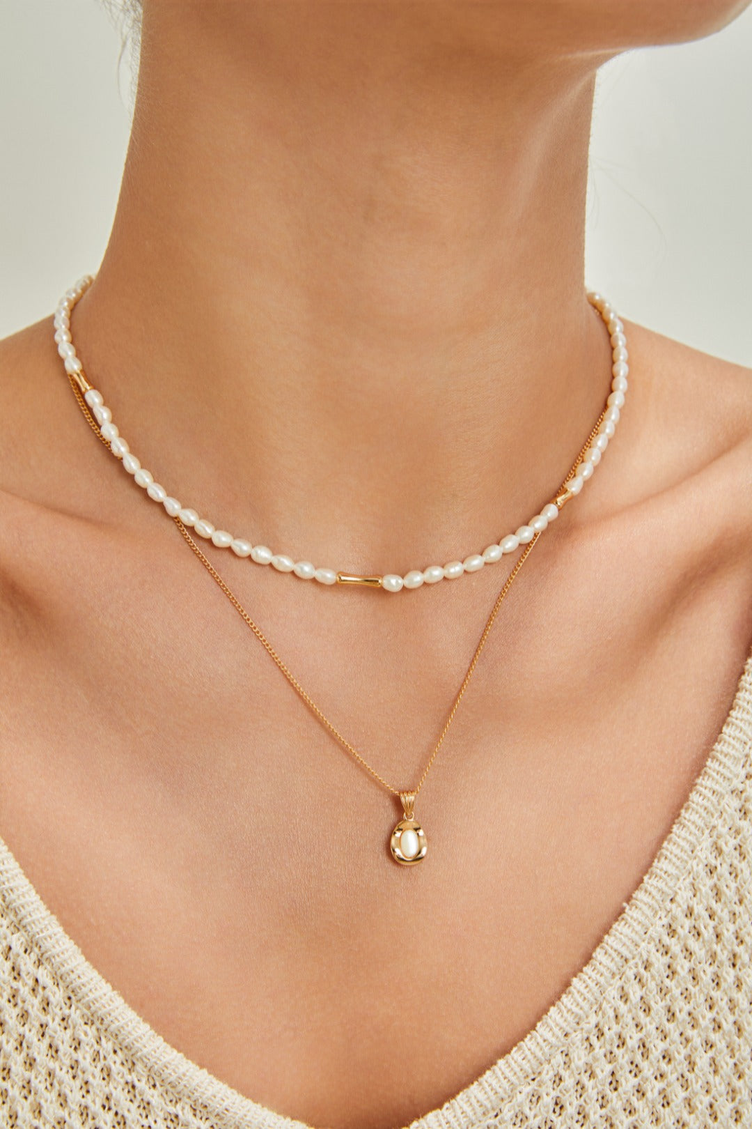 Close-up of a person wearing two necklaces. One is a short, delicate strand of small pearls, and the other is the Elegant Gold-Plated Sterling Silver Necklace with Teardrop Pendant by Siren Mode. The necklace features an adjustable length and a teardrop-shaped pendant. The person is wearing a cream-knit top, and the background is neutral.