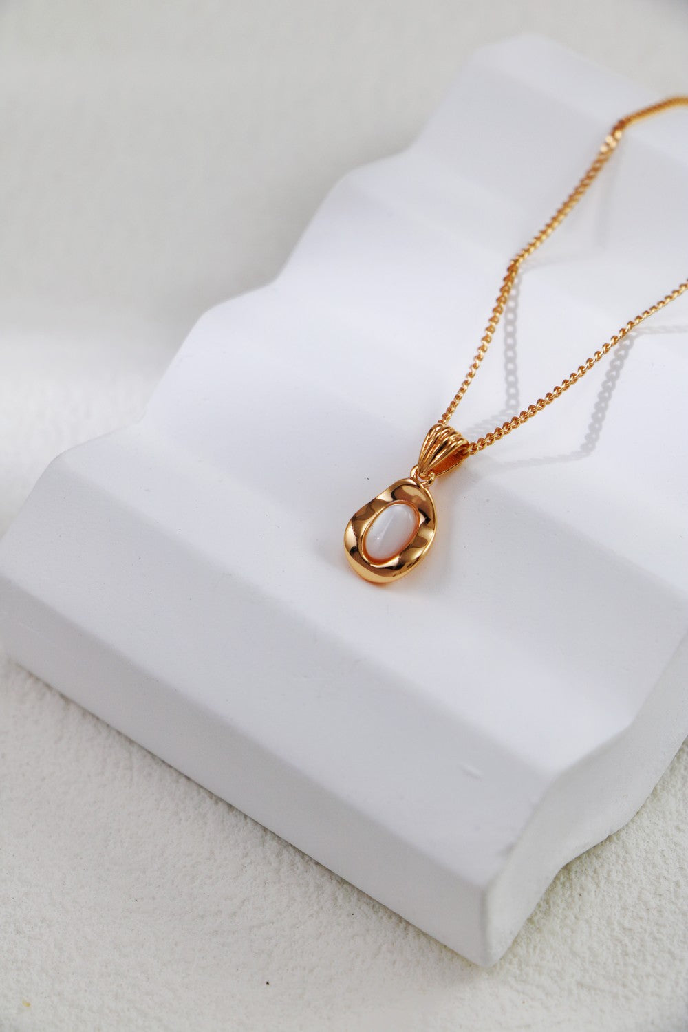 An Elegant Gold-Plated Sterling Silver Necklace with a teardrop pendant from Siren Mode is gracefully showcased on a white sculpted stand. The softly textured background accentuates the design's delicate and refined appearance.